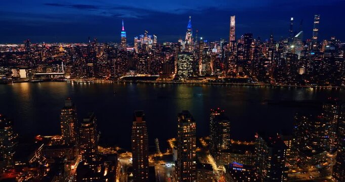 Descending over the luminous lively cityscape at night. View on the iconic skyline of Manhattan across the river with neon tops of skyscrapers standing out. New York City, USA.