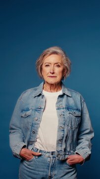 Confident senior woman with hands in pockets on blue backdrop. Stylish retiree poses in jeans and white t-shirt, embracing individuality and mature beauty.