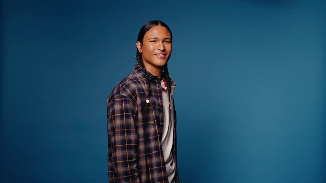 Young man with braids smiles at the camera in a medium shot against a blue backdrop. He wears a plaid shirt and a white t-shirt.