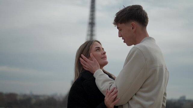 A young couple embraces affectionately in Paris France with the Eiffel Tower visible in the background The man and woman share a tender moment on an overcast day Medium shot handheld style.