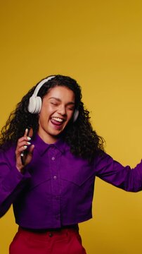Happy young woman with curly hair and headphones dances and sings against yellow background. Vertical medium shot.