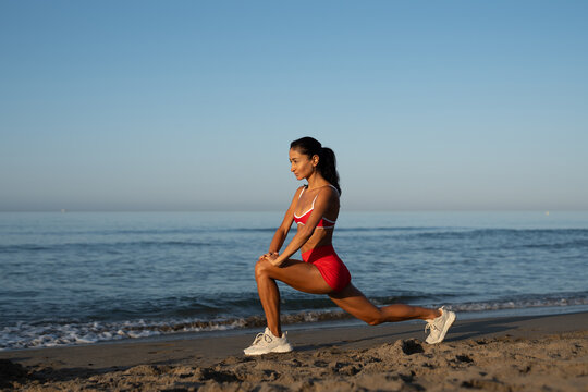 Focused athletic woman in red sportswear doing deep lunge stretch on sandy beach at sunrise, facing sideways toward ocean, enjoying peaceful morning workout by the sea
