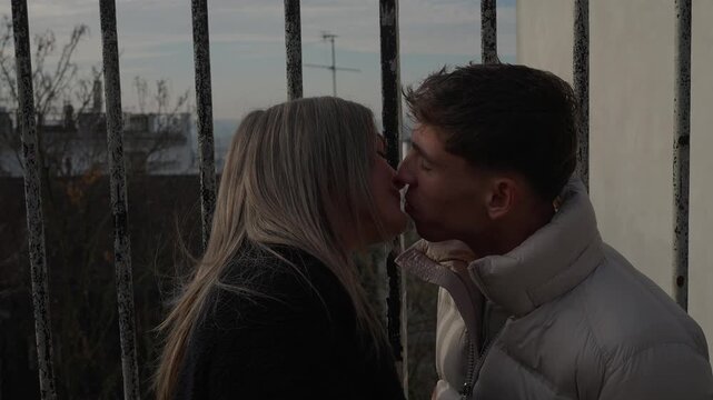 A young couple shares a romantic kiss in Paris France standing behind iron bars The handheld shot captures their love and affection in an urban setting highlighting the city's architecture and charm.