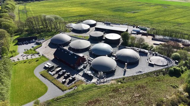 Close-up drone shot of circular anaerobic digesters at a biogas facility surrounded by yellow blooming rapeseed fields under bright sunlight.
