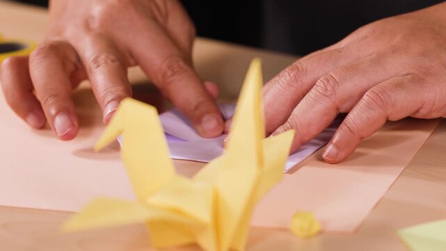 Hands folding purple paper into origami swan on wooden table with yellow paper crane.