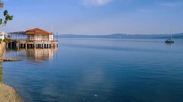 A charming restaurant perches on a pontoon, reflected in azure waters. A sailboat glides across the horizon on Evia island, Greece, where Mediterranean tranquility and beauty create a timeless escape