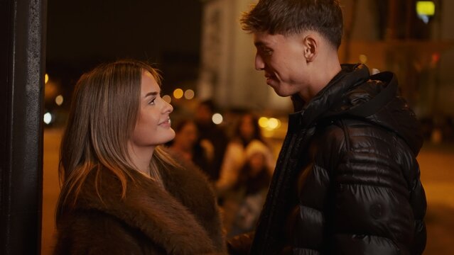 A romantic couple in Paris at night are looking at each other with love and affection wearing winter coats with city lights in the background. This image can be used for travel tourism or relationship