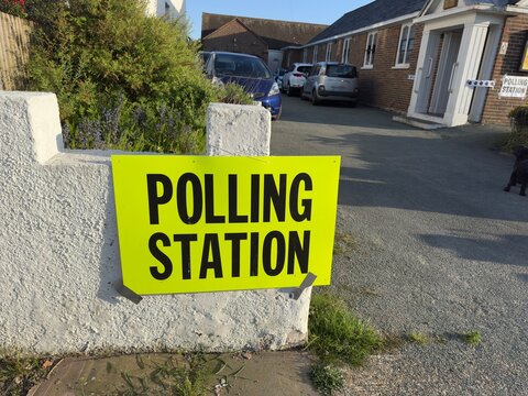 A luminous yellow sign with large black capital letters says ' POLLING STATION' indicating where people should vote in local and national elections. Placed on a street wall. Buildings in background.