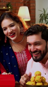 Friends video calling on Diwali festival, Indian Asian young couple friends in festive attire celebrating Hindu Deepavali at decorated home holding laddu barfi while attending video call on laptop