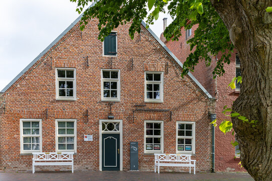 View of a traditional red brick building with white-framed windows and benches under a leafy tree in Wittmund, Lower Saxony, Germany.