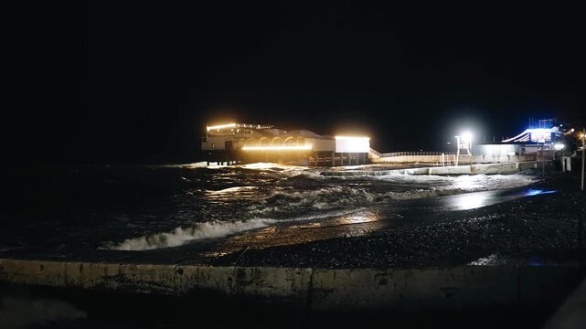 On a night beach by the sea, the lights on the shore are visible and the waves are visible.