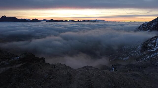 Aerial flyover along a ridge where hikers are enjoying the sunrise glow over the inversion of clouds in the Swiss Alps with a panoramic view of the mountains and colorful sky at dawn