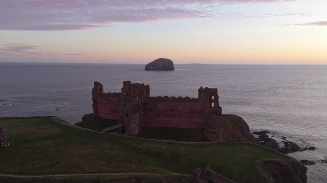 Aerial view tracking past the historic tantallon castle on the cliffs of east lothian, revealing bass rock in the north sea at dawn