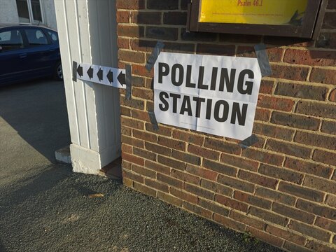 A white sign with large black capital letters says ' POLLING STATION' indicating where people should vote in local and national elections. Black arrows on white also point to where station is.