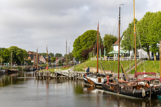View of Hafen Harlesiel with traditional sailing boats moored along the canal, surrounded by lush green trees and historic buildings in Wittmund, Lower Saxony, Germany.