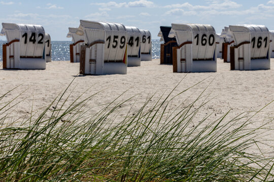 View of Strand Schillig with numbered traditional beach chairs on the sandy shore and green dune grass under a cloudy sky in Wangerland, Niedersachsen, Germany.