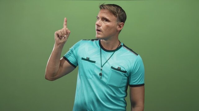 Man referee points finger upward with whistle on lanyard, wearing turquoise referee jersey and visible forearm against green studio backdrop; decisive call.