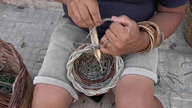 Male artisan weaving a basket with natural materials, using a wooden tool to shape the fibers, seated on a wooden platform surrounded by woven baskets