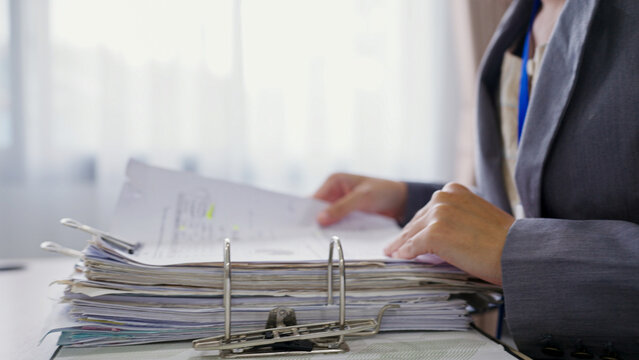 Woman reviews documents in a cluttered office environment. She focuses intently on paperwork with her fingers poised. The desk holds stacks of files and folders ready for sorting