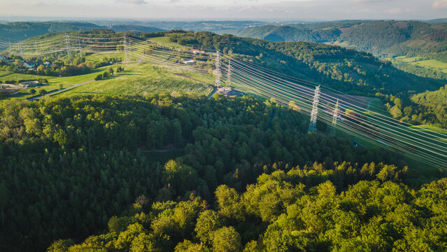 Aerial view of high voltage power lines crossing green forest and hills in Germany, energy transmission infrastructure in rural landscape at sunset