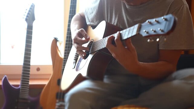 A man plays an acoustic guitar, focused on the strings. Electric guitars are visible in the background, creating a musical atmosphere in the room.