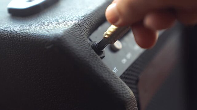 A hand is connecting a plug into the output of a guitar amplifier. The guitar amplifier has a textured surface and various control knobs visible.