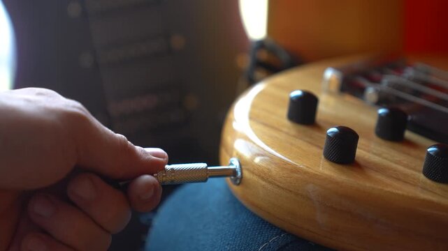 A man is connecting a cable to an electric guitar. The electric guitar has a smooth wooden finish and several control knobs visible in the scene.