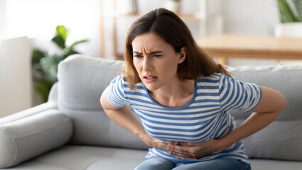 Obraz premium A woman experiencing stomach pain sits on a gray couch, clutching her abdomen in distress.