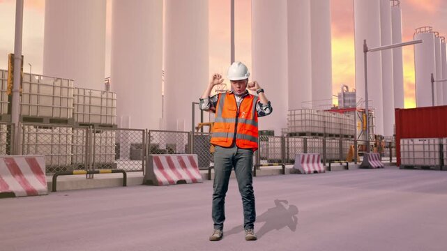 Full Body Of Asian Male Engineer With Safety Helmet Screaming Goal And Dancing Celebrating at Storage Facility with IBC Totes, Large Silos at Sunset