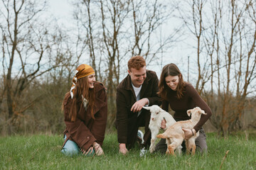 Fototapeta premium Lifestyle scene with three friends outdoors in a grassy field, interacting with playful goat kids, smiling and guiding the animals, capturing a warm candid moment of friendship and nature.