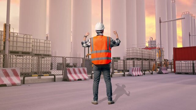 Full Body Back View Of A Male Engineer With Safety Helmet Raising His Hands Celebrating While Working at Storage Facility with IBC Totes, Large Silos at Sunset
