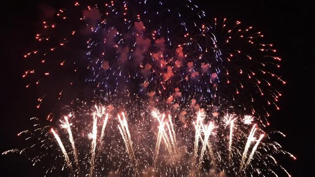 Festive display of vibrant red, white, and blue fireworks exploding in the dark night sky with thick smoke clouds and shimmering light trails during a pyrotechnic holiday celebration event.