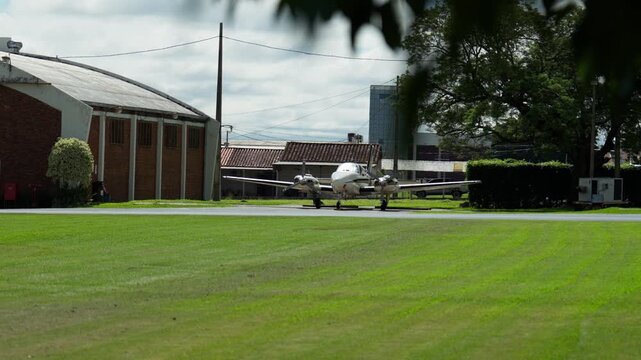 Small twin engine propeller airplane parked near hangar buildings on a green grass airfield. Shows private aviation, general aviation setting, and aircraft positioned on the ground.