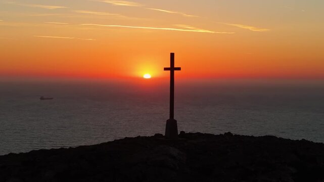 A cross stands silhouetted against a vibrant sunrise over the ocean, creating a serene and contemplative scene. Ideal for spiritual or travel content.