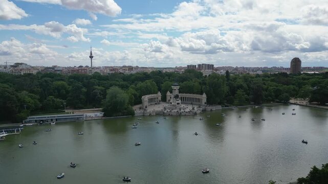 The Monument to Alfonso XII surrounded by boats on a lake in El Retiro Park. The lush greenery complements the cityscape in the background. Suitable for travel and urban content.