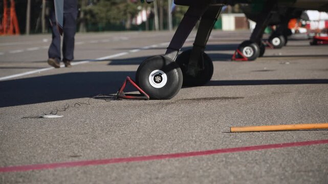 Wheels of light aircraft with chocks stand on airport ramp on sunny day. In background of parked aircraft along marking legs of walking mechanic are visible.