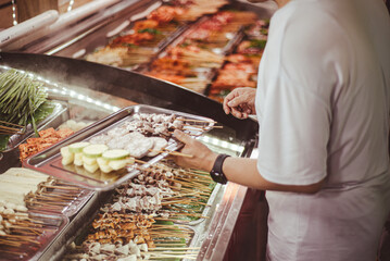 Obraz premium Chilled skewer display features lotus root, zucchini, tofu, and assorted meats arranged in refrigerated trays while a shopper builds a tray of chosen items, Mengzi, Yunnan