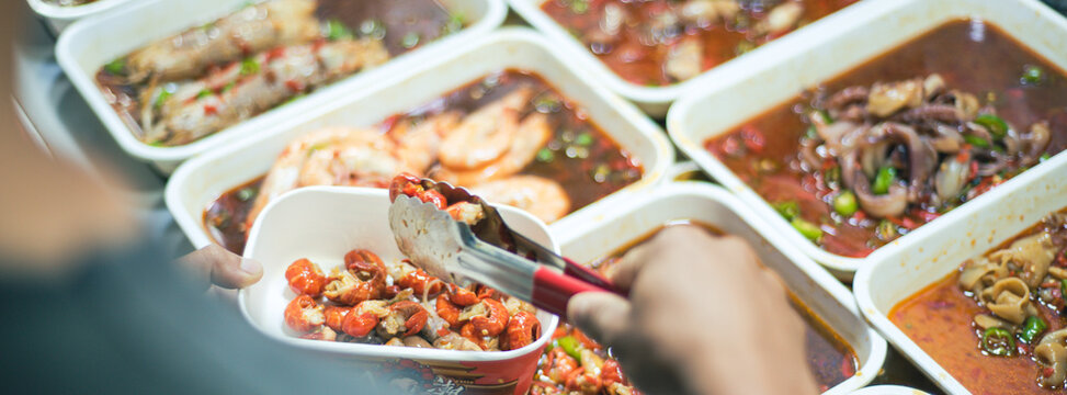 Vendor ladling chili broth lifts seasoned shrimp and squid from a heated tray infused with peppers and aromatic oils under night market glow, Mengzi, Yunnan