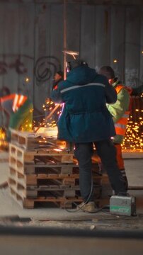 At vacant construction site, builders protective gear wield metal cutters, shaping steel beams as sparks fly. Wooden pallets half-built frames surround them, highlighting their focused craftsmanship