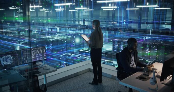 IT Specialist Types at a Workstation While a Female Analyst Uses Tablet. Luminous Lines of Information Overlays Visualize Network Flow, Server Activity, System Working.