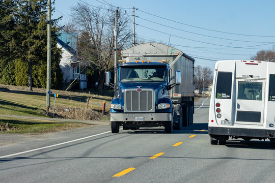 Quebec, Canada, 24 April 2026 : A blue Peterbilt semi truck with a trailer drives on a rural road past a white bus
