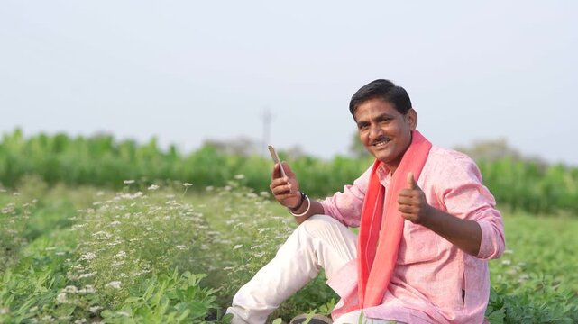 young indian farmer using smartphone sitting at green agricultural field