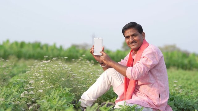 young indian farmer showing smartphone screen sitting at green agricultural field