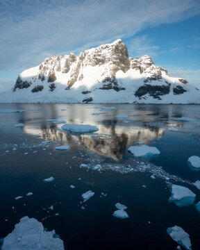 View of a massive snow-covered mountain peak reflecting in the calm dark blue waters filled with floating icebergs and ice floes under a clear sky in Antarctica.