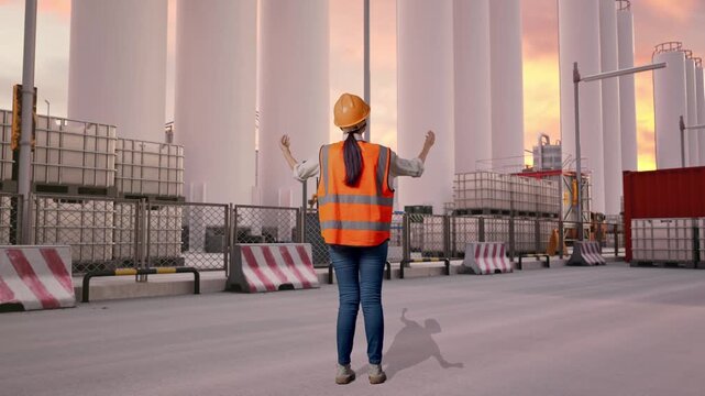 Full Body Back View Of A Female Engineer With Safety Helmet Spreading Arms While Standing at Storage Facility with IBC Totes, Large Silos at Sunset