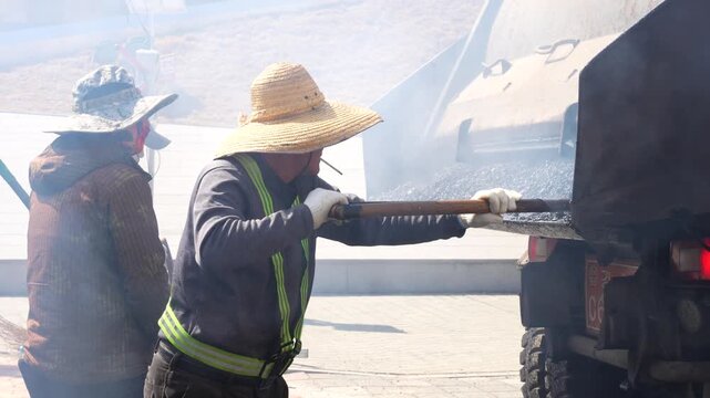 Korean road workers with shovels unload hot smoking asphalt from a dump truck for paving a new street