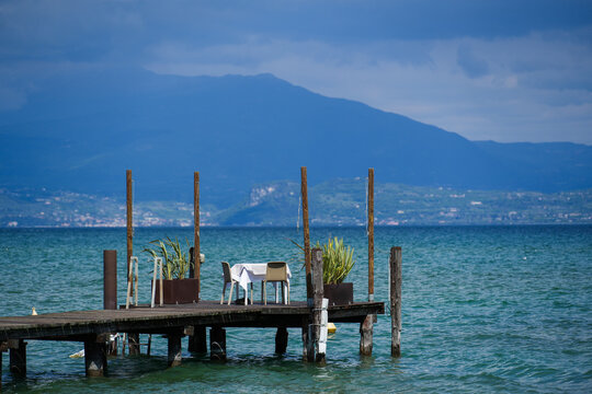 View of a table for two set on a wooden pier overlooking the blue waters of Lake Garda with mountains in the background Sirmione, Lombardy, Italy.