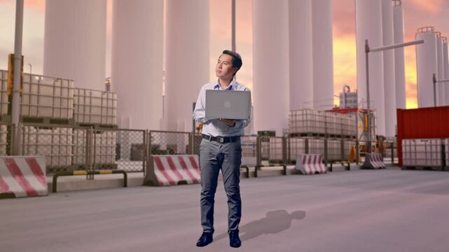 Full Body Of An Asian Male Professional Worker With His Laptop at Storage Facility with IBC Totes, Large Silos at Sunset, He Observes By Looking Around Before He Come To Concentrating With His Laptop