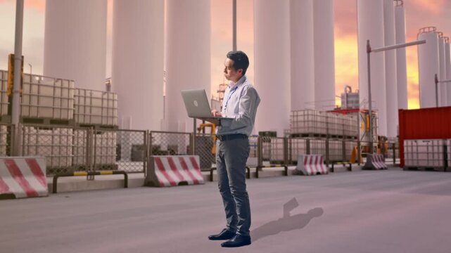 Full Body Side View Of An Asian Male Professional Worker Standing With His Laptop at Storage Facility with IBC Totes, Large Silos at Sunset, Typing And Dissapionted With Nodding His Head 