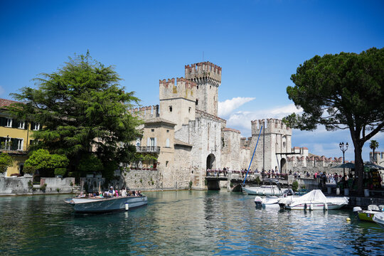 View of the medieval Scaligero Castle with its stone towers and bridge over the turquoise water of Lake Garda with tourists and boats Sirmione, Lombardy, Italy.